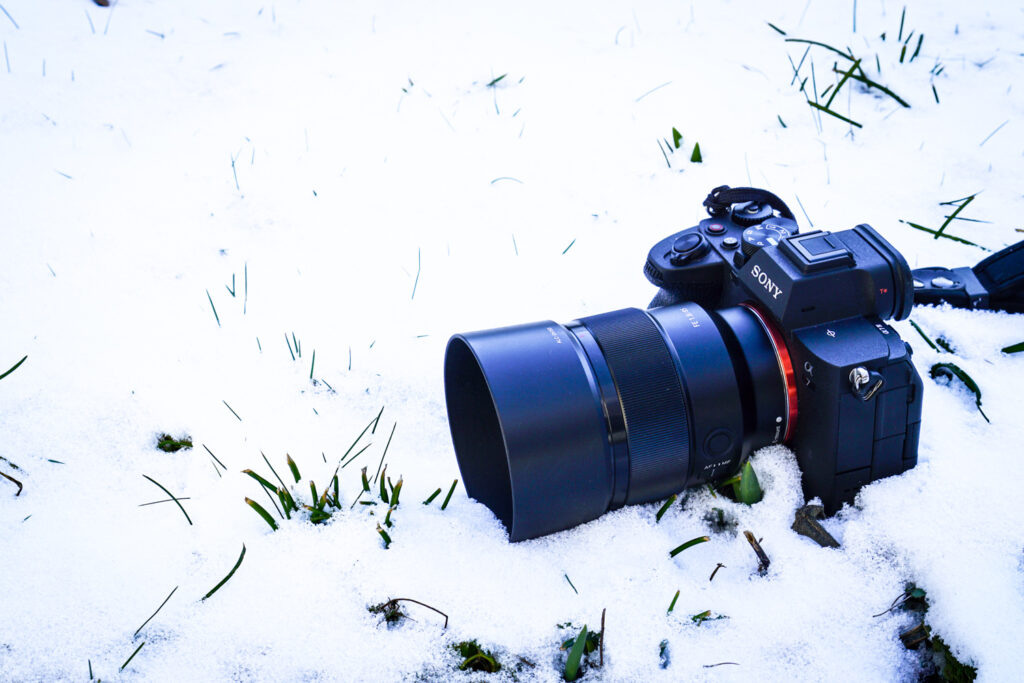 Spiegellose Kamera mit Objektiv liegt im Schnee, umgeben von hervorschauendem Gras