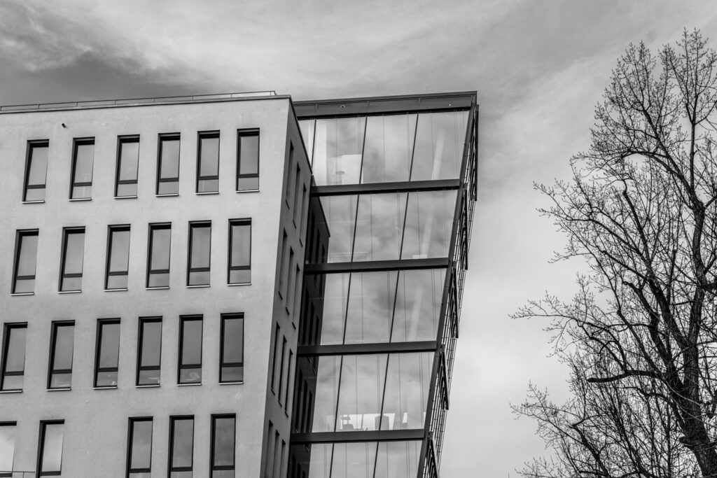 Schwarzweisse Aufnahme eines modernen Gebaeudes mit Glasfassade und klaren Linien, daneben ein kahler Baum.