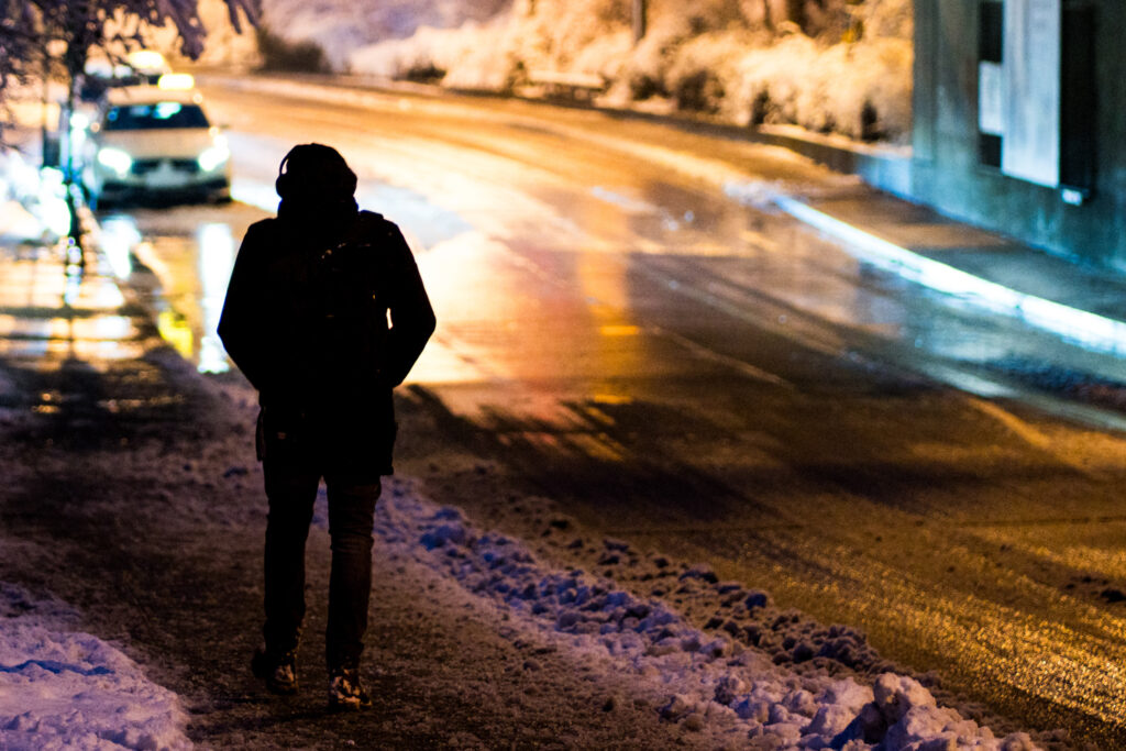 Silhouette einer Person mit Rucksack geht nachts durch verschneite, beleuchtete Straße