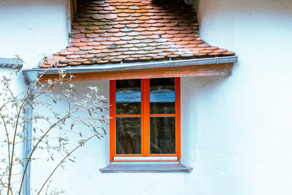 Orange gerahmtes Fenster in heller Hauswand unter Dach mit runden Ziegeln