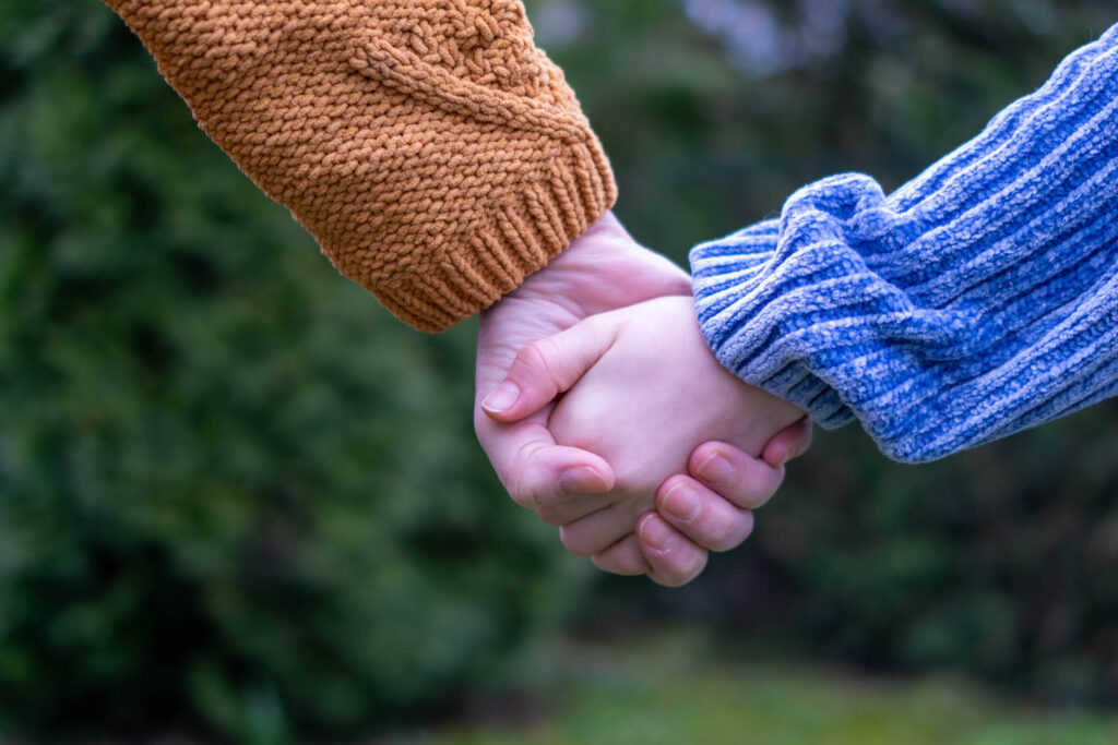 Nahaufnahme von zwei Personen, die sich an den Händen halten. Eine Person trägt einen orangefarbenen, grob gestrickten Pullover, die andere einen blau-weiß gestreiften Pullover aus weichem Stoff. Der Hintergrund ist unscharf und zeigt grüne Natur, vermutlich Bäume oder Sträucher.
