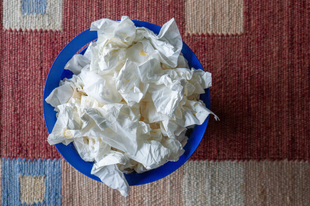 Ein blauer Papierkorb, der mit vielen zerknüllten weißen Papiertüchern gefüllt ist. Der Korb steht auf einem Teppich mit rotem und blauem geometrischen Muster.