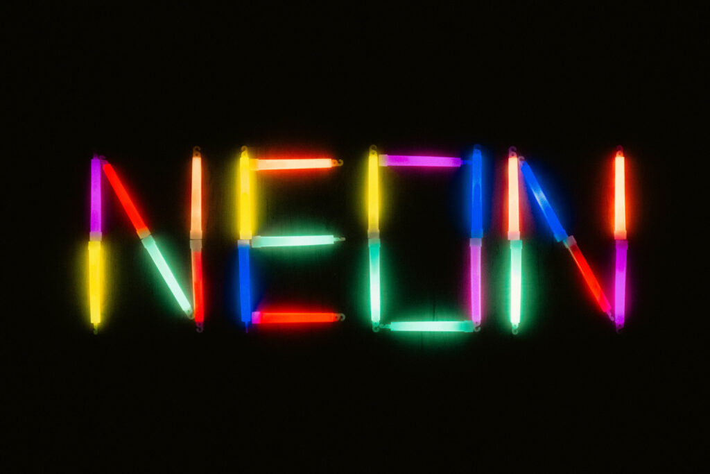Das Wort 'NEON' ist aus leuchtenden, bunten Neonröhren auf einem dunklen Hintergrund geformt. Die Röhren strahlen in verschiedenen Farben wie Rot, Gelb, Blau, Grün und Lila und erzeugen einen lebendigen, leuchtenden Effekt.