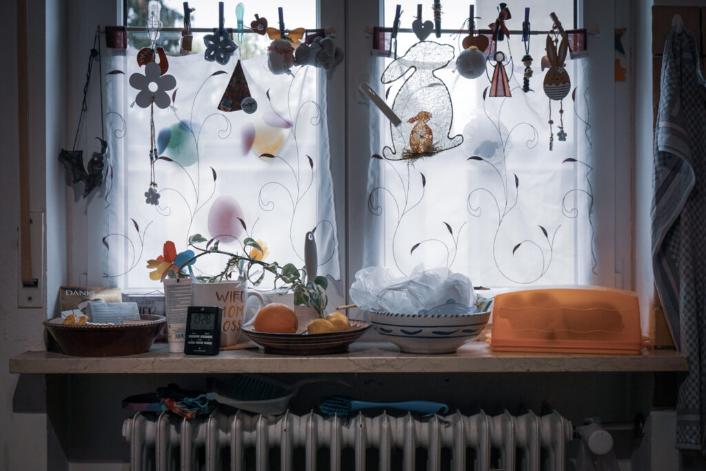 Ein Fensterbrett in einer Küche ist dekoriert mit österlichen und frühlingshaften Gegenständen. An der Gardinenstange hängen diverse bunte Anhänger, darunter Osterhasen, Blumen und Eier. Die weißen Gardinen sind mit floralen Mustern verziert. Auf dem Fensterbrett stehen verschiedene Schalen mit Obst, eine Tasse mit der Aufschrift „WIFE MOM BOSS“, ein digitaler Wecker, eine Pflanze, eine orangefarbene Tortenhaube und Haushaltsgegenstände. Unter dem Fensterbrett befindet sich ein Heizkörper, über dem einige Reinigungsutensilien hängen. Die Szene wirkt gemütlich und liebevoll dekoriert.