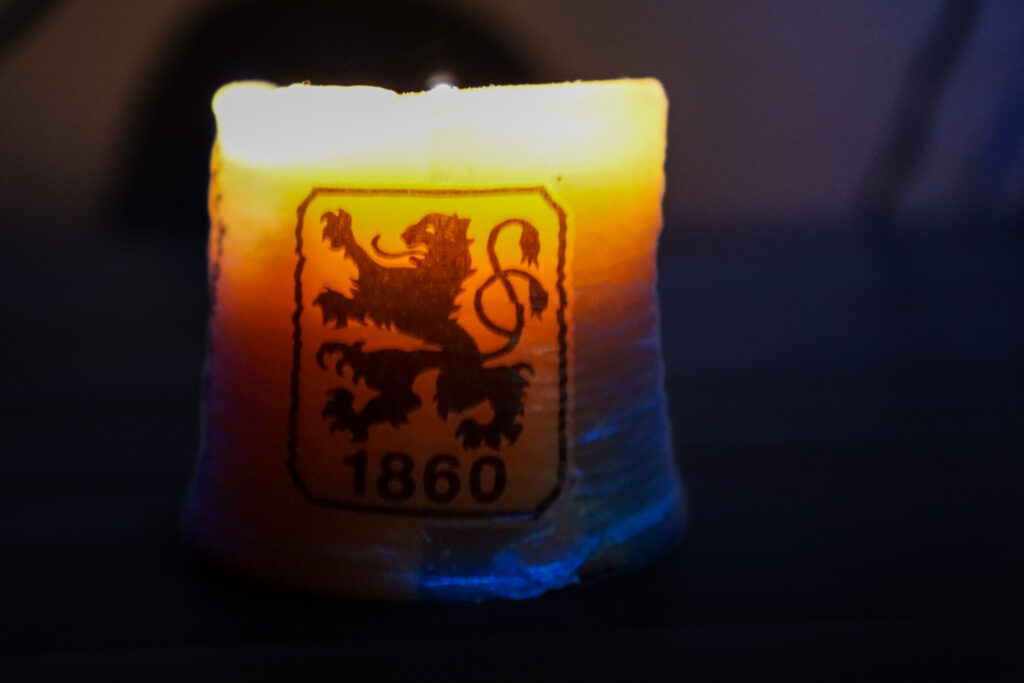 Eine brennende Kerze mit einem Wappenaufdruck, der einen aufrecht stehenden Löwen und die Zahl '1860' zeigt. Die Kerze leuchtet warm in Gelb- und Orangetönen, der Hintergrund ist dunkel und unscharf.