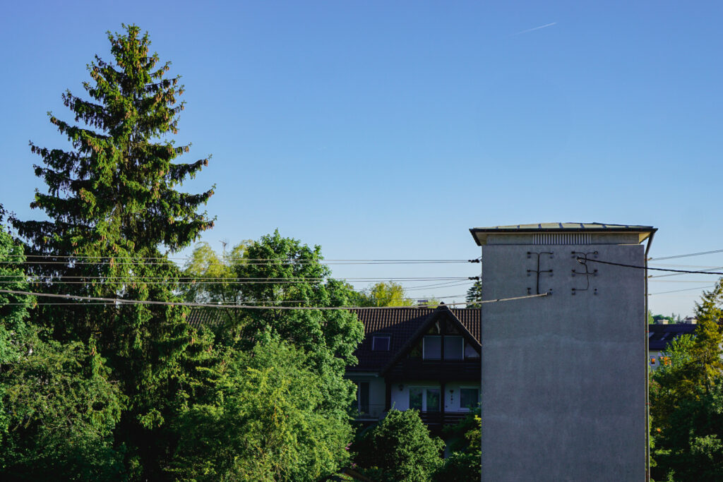 Das Bild zeigt eine Freileitung mit mehreren Stromkabeln, die an einem grauen, turmartigen Gebäude befestigt sind. Im Hintergrund befinden sich ein Wohnhaus mit dunklem Satteldach sowie zahlreiche grüne Bäume. Der Himmel ist klar und blau.