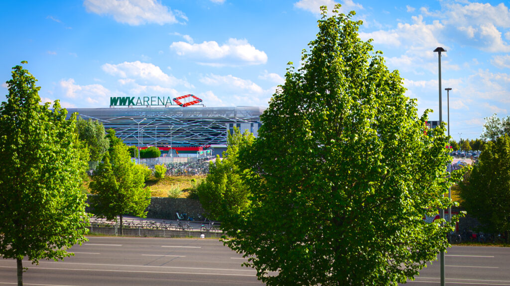 Blick auf die WWK Arena hinter grünen Bäumen an einer breiten Strasse unter blauem Himmel mit Wolken.