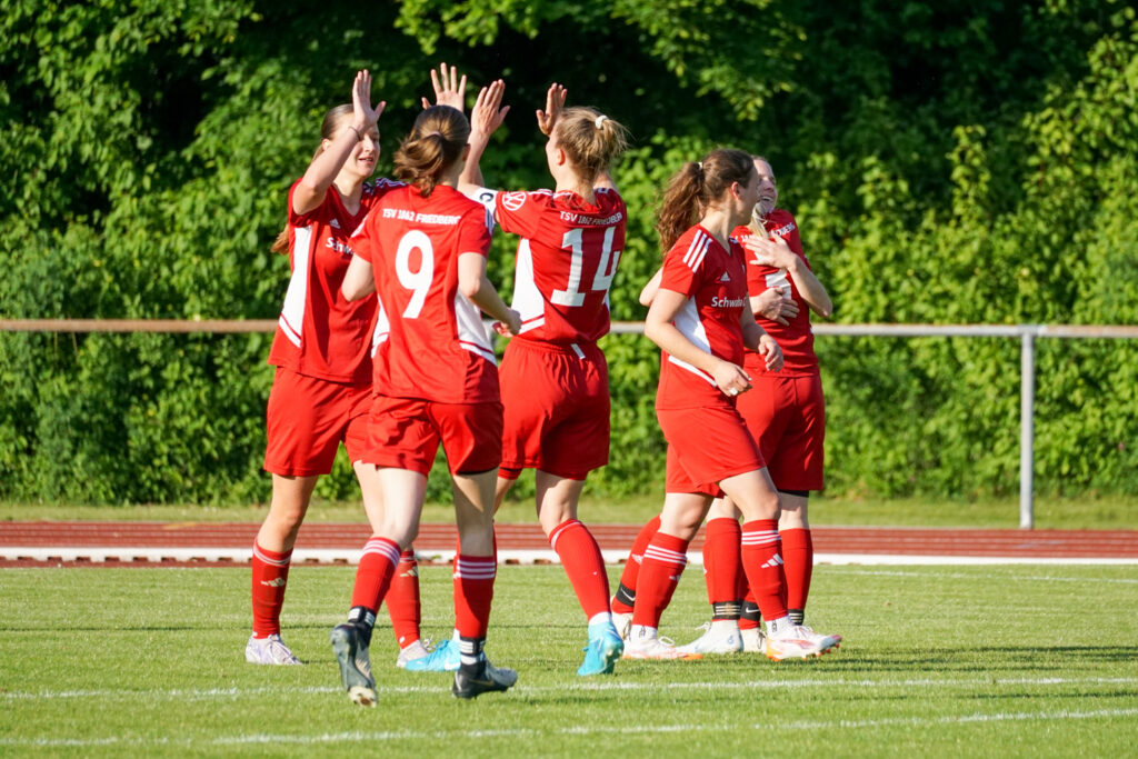 Eine Gruppe von Fußballspielerinnen in roten Trikots feiert gemeinsam auf dem Spielfeld, vermutlich nach einem Tor. Im Hintergrund ist eine grüne Hecke zu sehen.