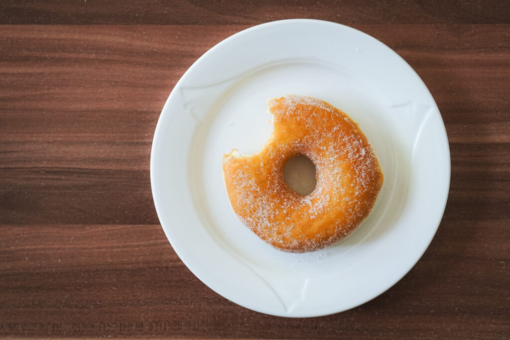 Das Bild zeigt einen zuckerbestreuten Donut mit einem Biss, der auf einem weißen Teller liegt. Der Teller steht auf einer Holzoberfläche mit einer dunkelbraunen Maserung. Der Donut ist goldbraun und hat die typische Ringform, wobei der Zucker gut sichtbar auf der Oberfläche verteilt ist.