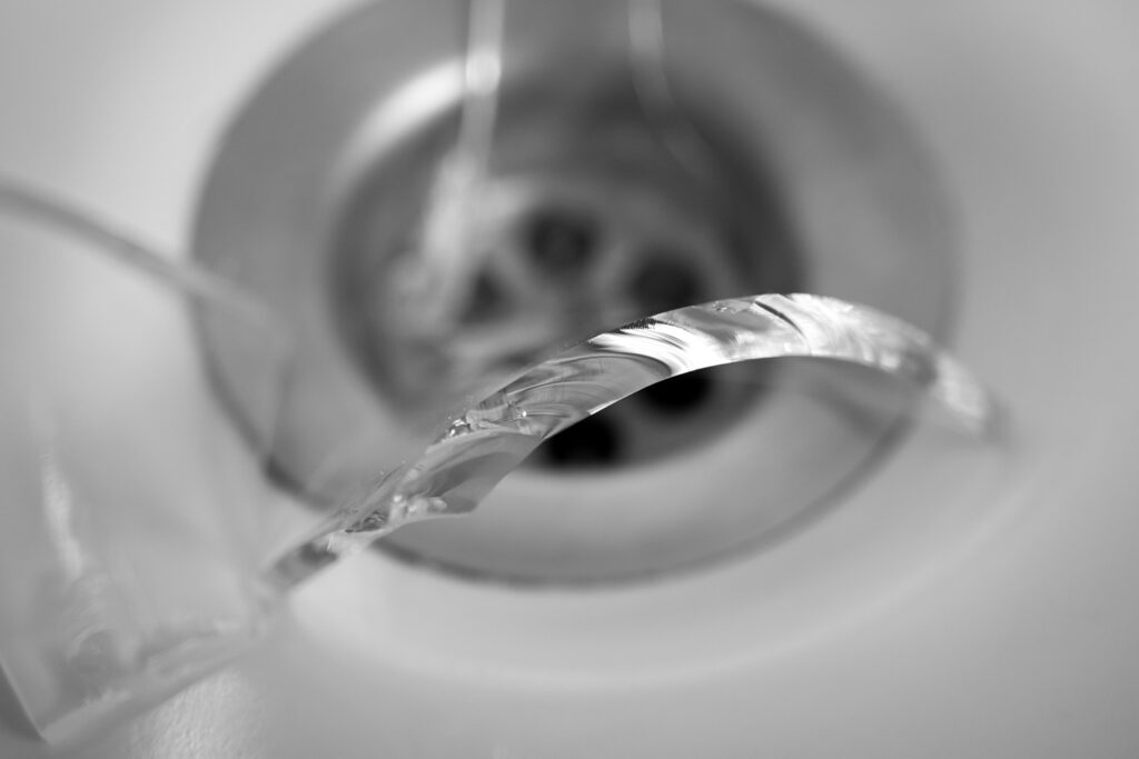 Nahaufnahme eines zerbrochenen Glasrandes über einem Abfluss in einem Waschbecken, in Schwarz-Weiß fotografiert.