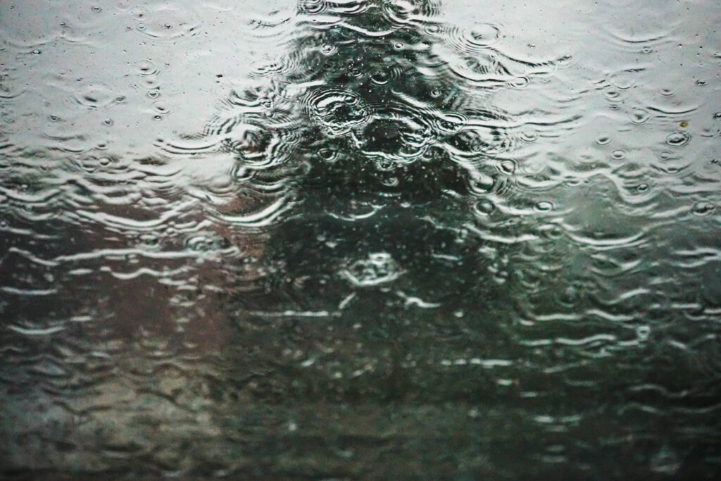 Regentropfen auf einer Fensterscheibe mit kreisförmigen Wellen und verschwommenem Baum im Hintergrund.