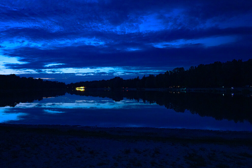 Blaue Abendstimmung an einem See mit spiegelglatter Wasseroberfläche, dunklen Bäumen am Ufer und einem hell erleuchteten Gebäude in der Ferne.