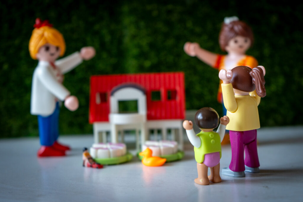 Vier Playmobil-Figuren stellen eine Familie dar, bestehend aus zwei Erwachsenen und zwei Kindern, die sich vor einem kleinen weißen Spielzeughaus mit rotem Dach versammeln. Im Vordergrund stehen die Kinder mit erhobenen Armen, während die Erwachsenen im Hintergrund mit ausgestreckten Armen zu ihnen blicken. Vor dem Haus liegen zwei grüne Teller mit weißen Tortenstücken sowie eine kleine gelbe Plastikente auf dem Boden. Der Hintergrund ist unscharf und grün, was eine Hecke oder Pflanzen andeutet.