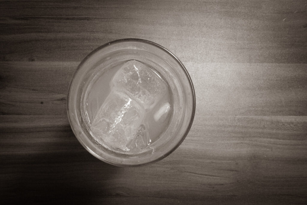Draufsicht auf ein Glas mit Eiswürfeln und einem klaren Getränk auf einer Holzoberfläche in Schwarz-Weiss.