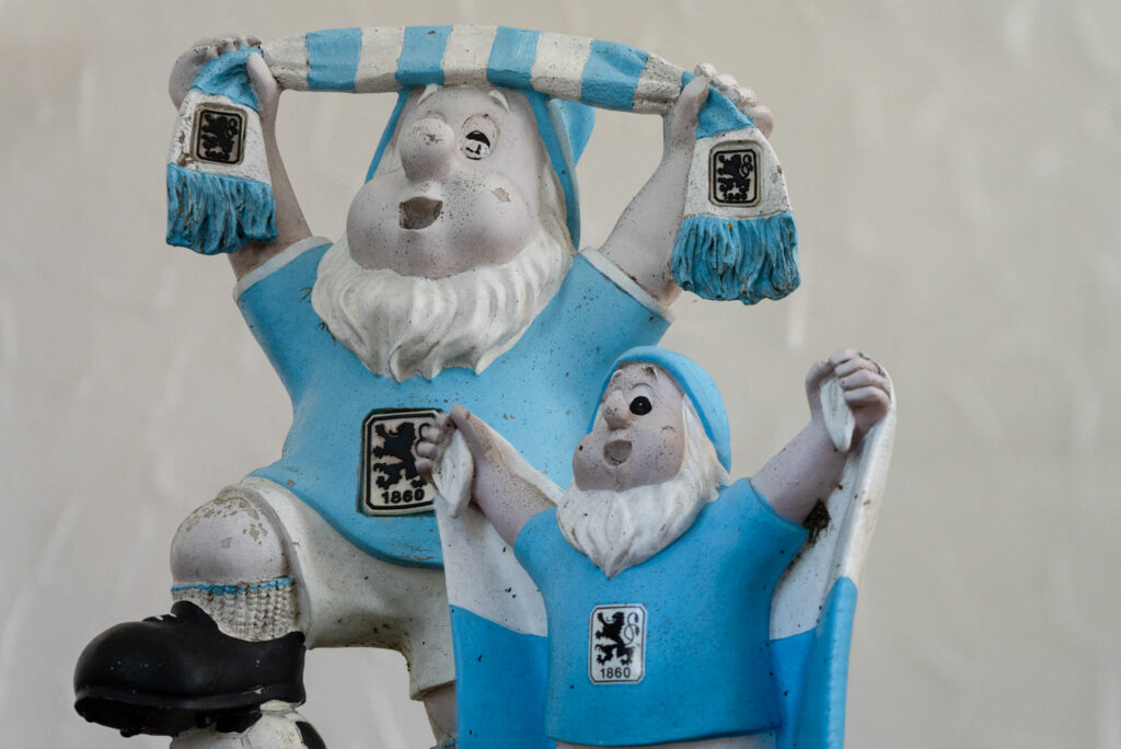 Zwei Figuren mit weißen Bärten in Fanbekleidung des TSV 1860 München. Die größere Figur trägt ein hellblaues Trikot und hält einen blau-weiß gestreiften Schal über dem Kopf. Die kleinere Figur daneben trägt ebenfalls ein hellblaues Trikot und trägt mit nach oben ausgestreckten Armen eine weiß-blaue Fahne hinter dem Rücken. Beide Figuren zeigen das Vereinswappen auf der Brust.