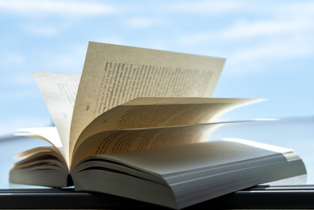 Ein aufgeschlagenes Buch liegt auf einer Fensterbank, während der Wind mehrere Seiten bewegt. Im Hintergrund ist ein blauer Himmel mit einigen Wolken zu sehen.