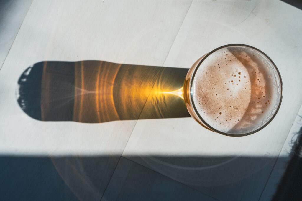 Ein Glas mit goldenem, schäumendem Getränk steht auf einer hellen Oberfläche im Sonnenlicht. Der Blick von oben zeigt den cremigen Schaum auf dem Getränk, während das Glas einen langen, warm leuchtenden Schatten wirft und das Licht in verschiedenen Goldtönen reflektiert.