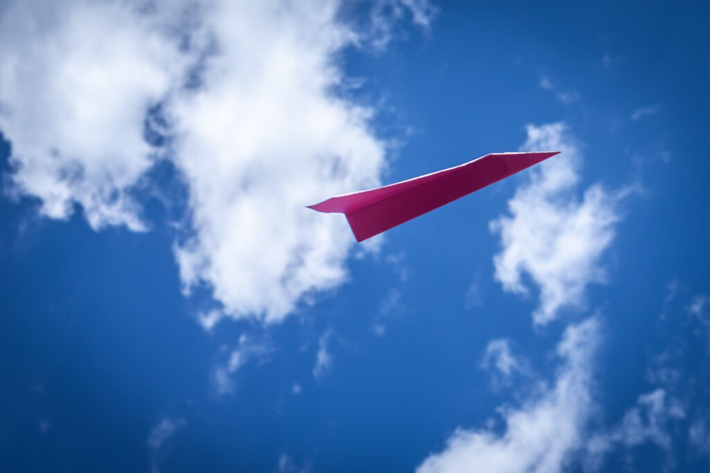 Rosaroter Papierflieger fliegt vor blauem Himmel mit weissen Wolken.