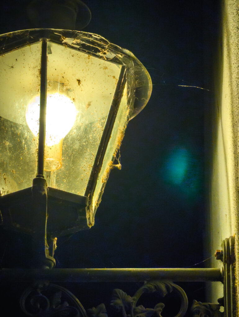 Nahaufnahme einer beleuchteten, verwitterten Aussenlampe mit Spinnweben vor dunklem Hintergrund.