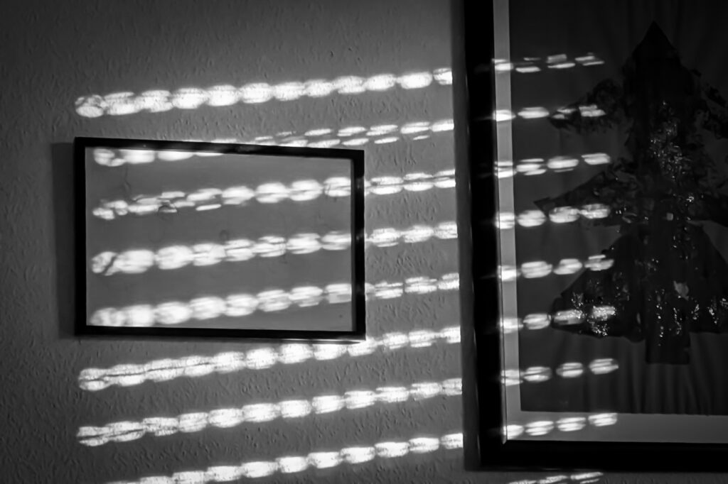 Schwarzweisse Wand mit Bilderrahmen, überzogen von leicht diagonal verlaufenden Lichtstreifen einer Jalousie.
